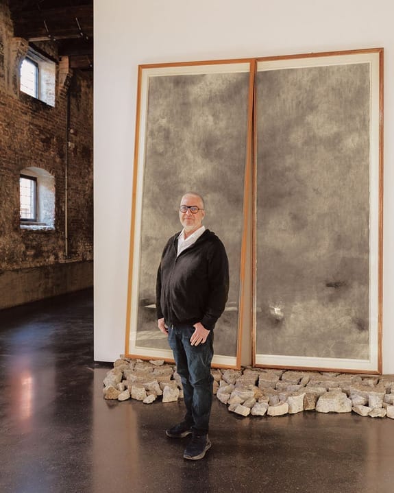 Person standing in front of large abstract gray artwork on display in an industrial-style gallery with brick walls.