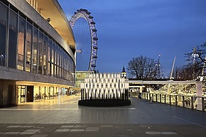 Illuminated modern art installation outside a building with the London Eye and Big Ben in the background at dusk.