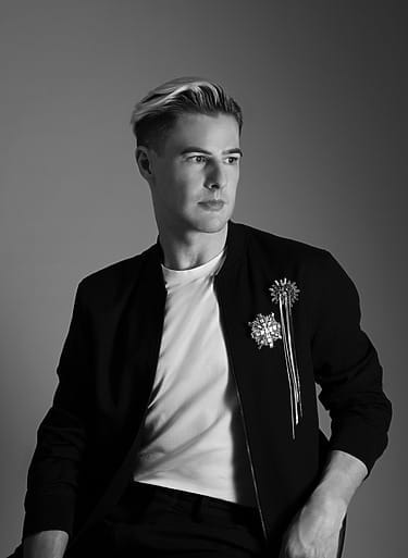 Black and white photo of a man in a stylish jacket with decorative brooches, looking to the side against a gray background.