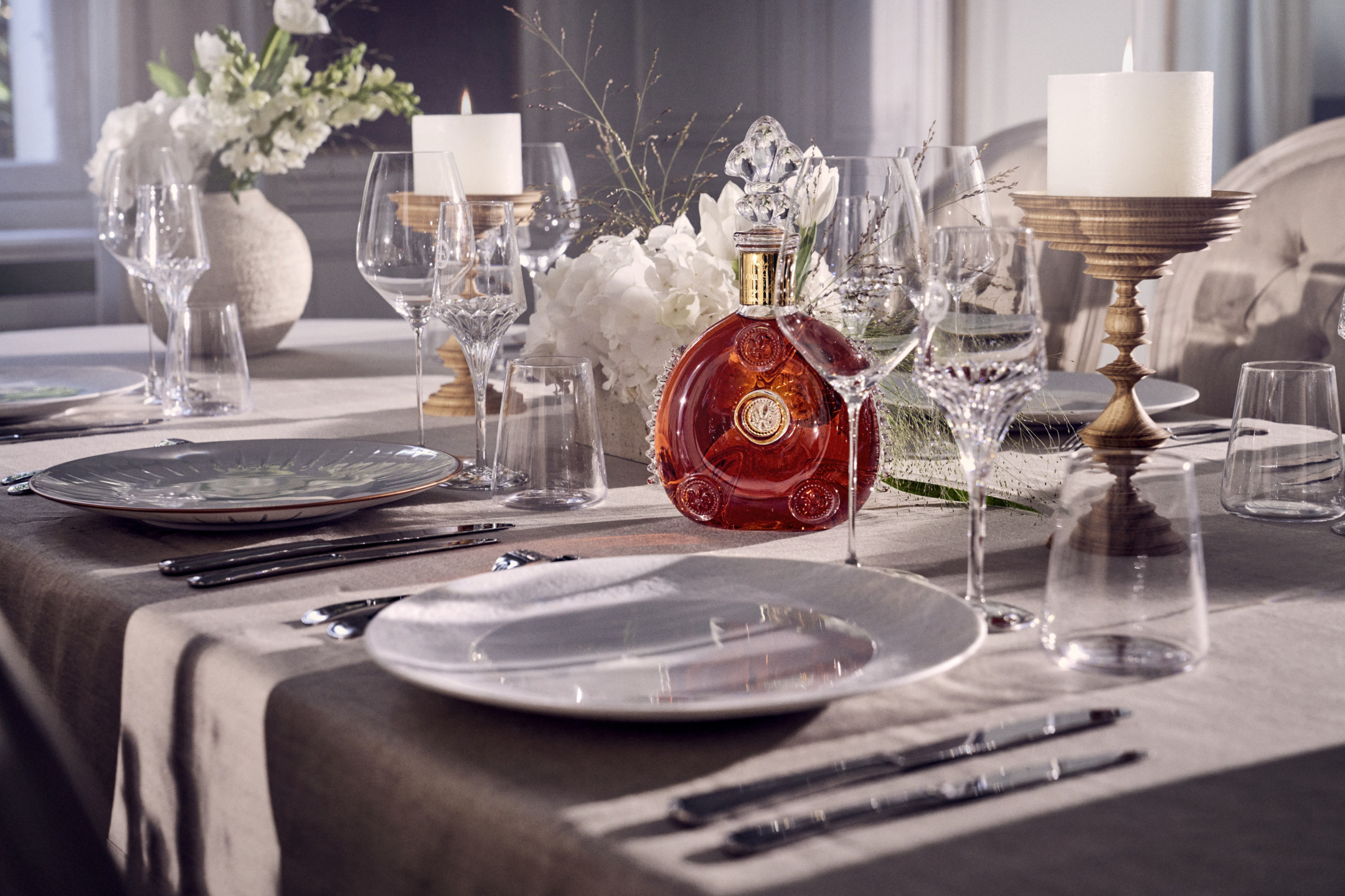 Elegant dining table with crystal glassware, a red bottled liquor, candles, and floral centerpiece in a cozy setting.
