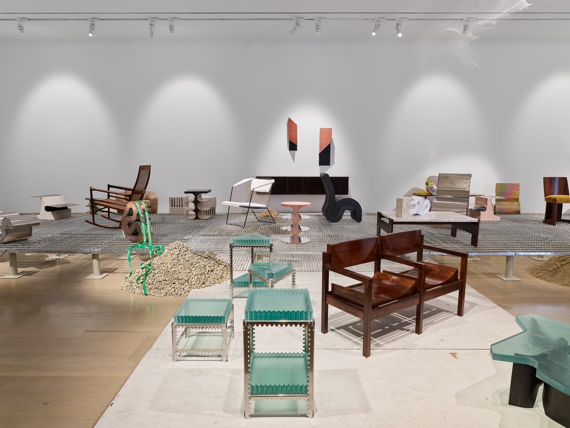Modern art exhibit showcasing various chairs and tables on a raised platform in a gallery setting with a white wall backdrop.