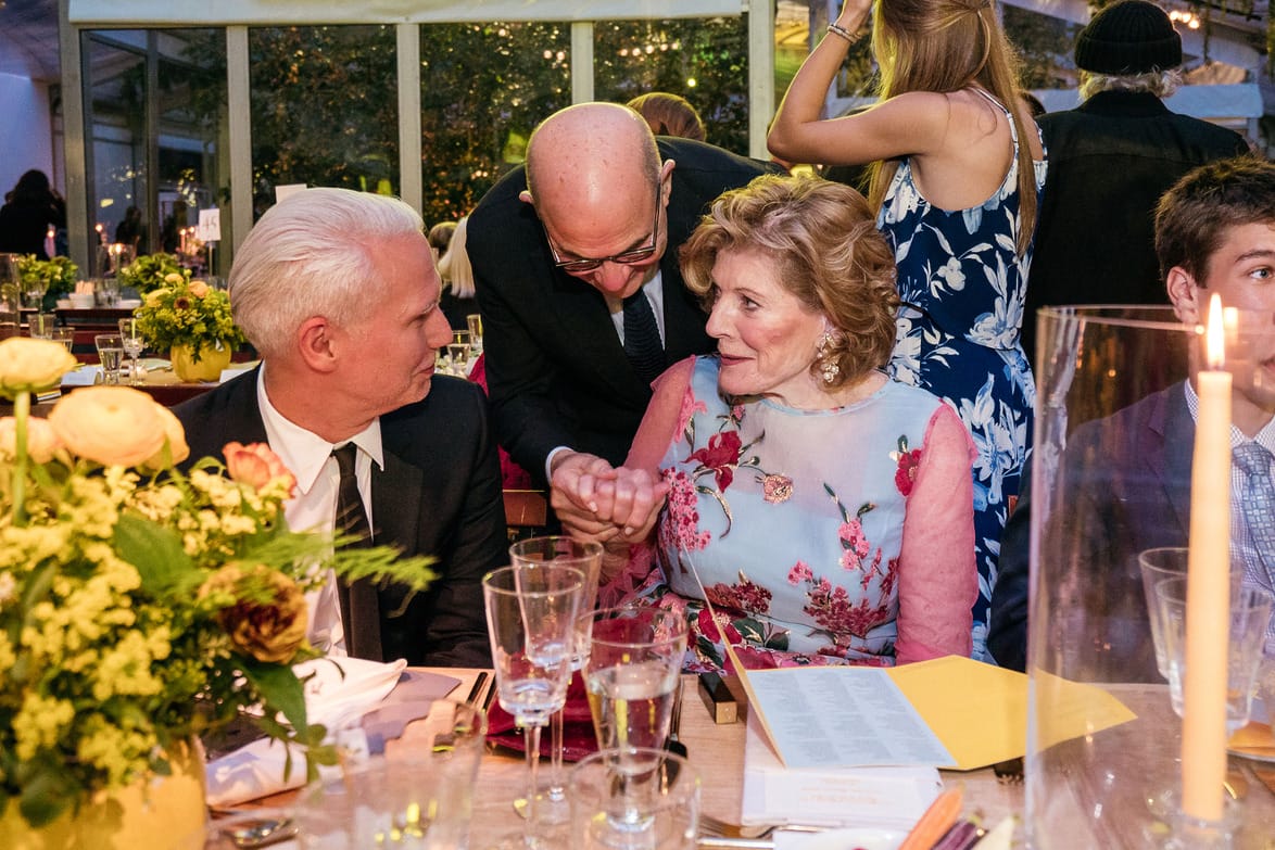 Group of people in formal attire talking and enjoying a social event at a decorated table with flowers and candles.