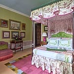 Colorful bedroom with a four-poster bed, floral canopy, vibrant decor, green walls, framed art, and a pink upholstered chair.