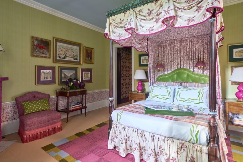 Colorful bedroom with a four-poster bed, floral canopy, vibrant decor, green walls, framed art, and a pink upholstered chair.