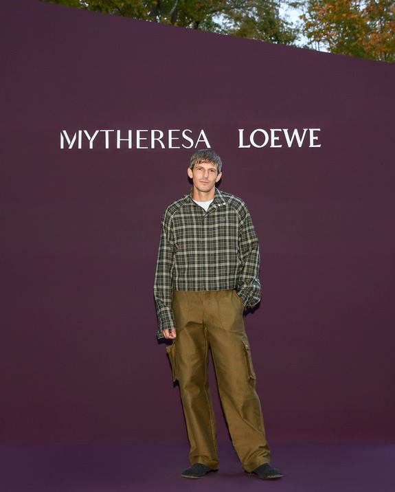 Person in checkered shirt and brown pants standing in front of Mytheresa and Loewe backdrop.
