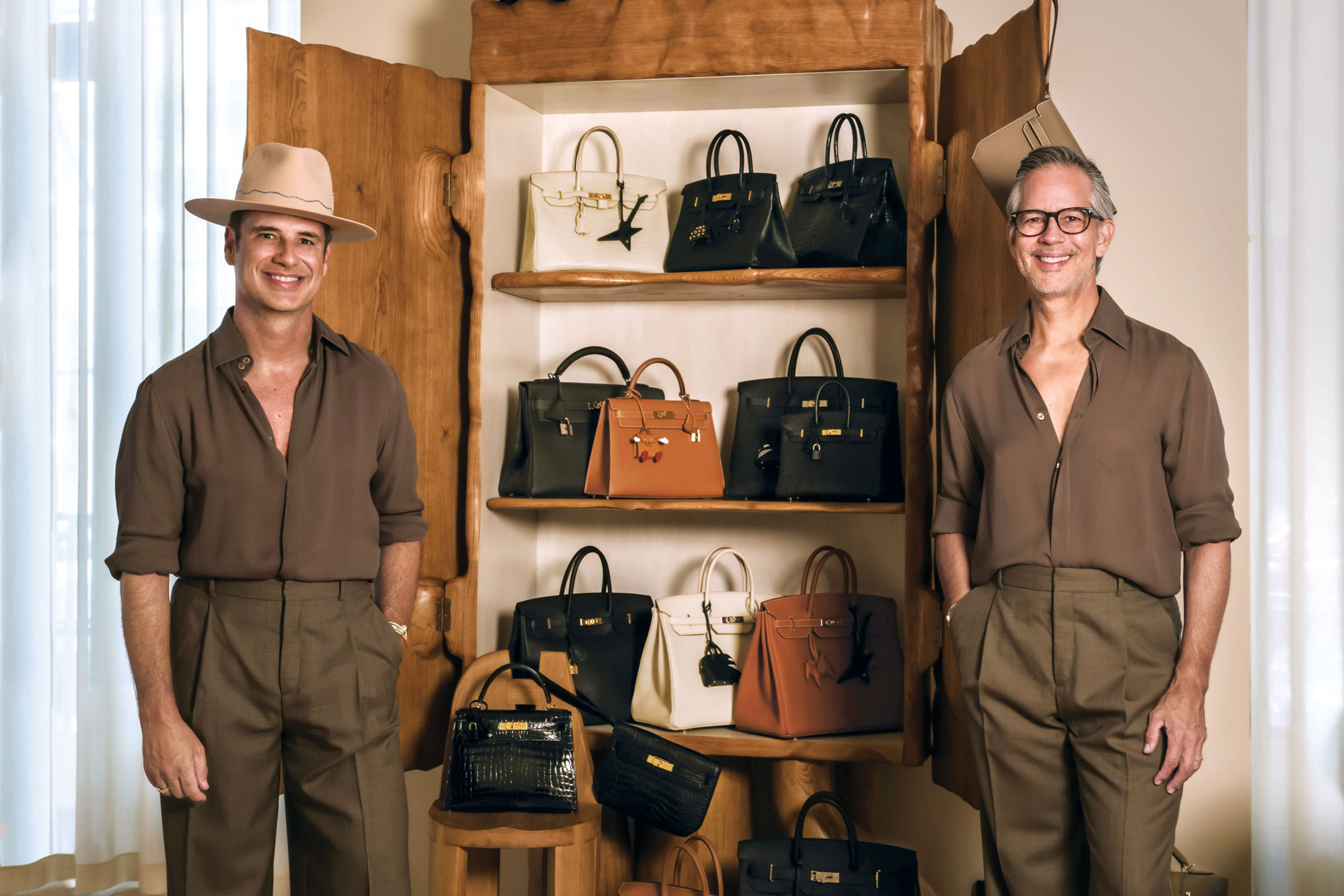 Two people standing by a wooden shelf displaying various luxury handbags, both wearing brown outfits and smiling.