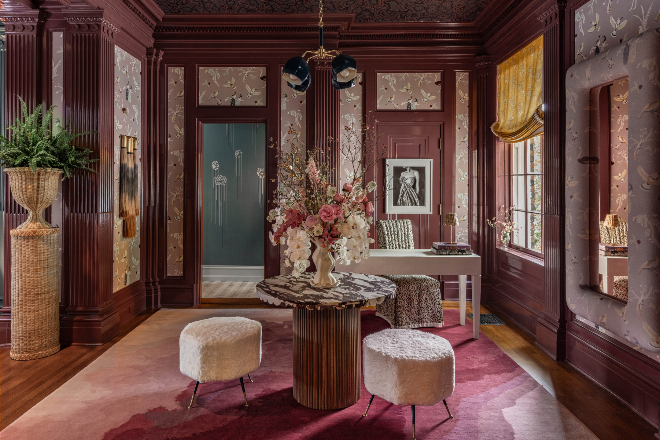 Elegant room with floral wallpaper, marble table, plush stools, and a large window with golden curtains.