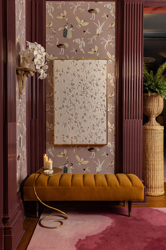 Elegant living room with floral wallpaper, gold frame art, mustard bench, candles, orchids, and woven vase with green plants.