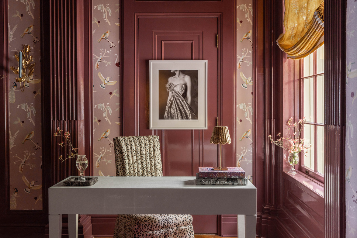 Elegant home office with pink walls, floral wallpaper, a white desk, patterned chair, small lamp, and framed black-and-white photo.