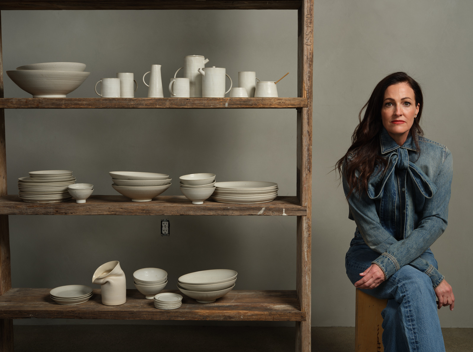 Woman in denim seated next to a wooden shelf displaying an array of white ceramic bowls and plates.