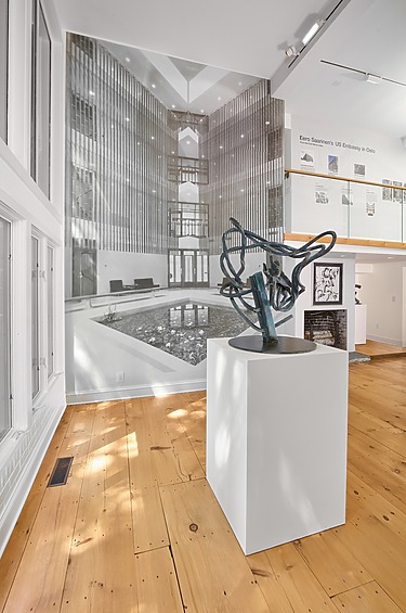 Art gallery interior with sculpture on a pedestal, large black and white wall photo, and wooden floor.