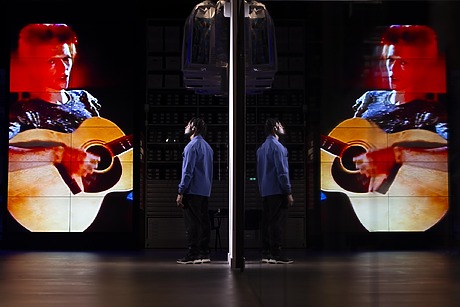 Person in blue looking at a large screen displaying a musician playing guitar, mirrored reflection on the surface.