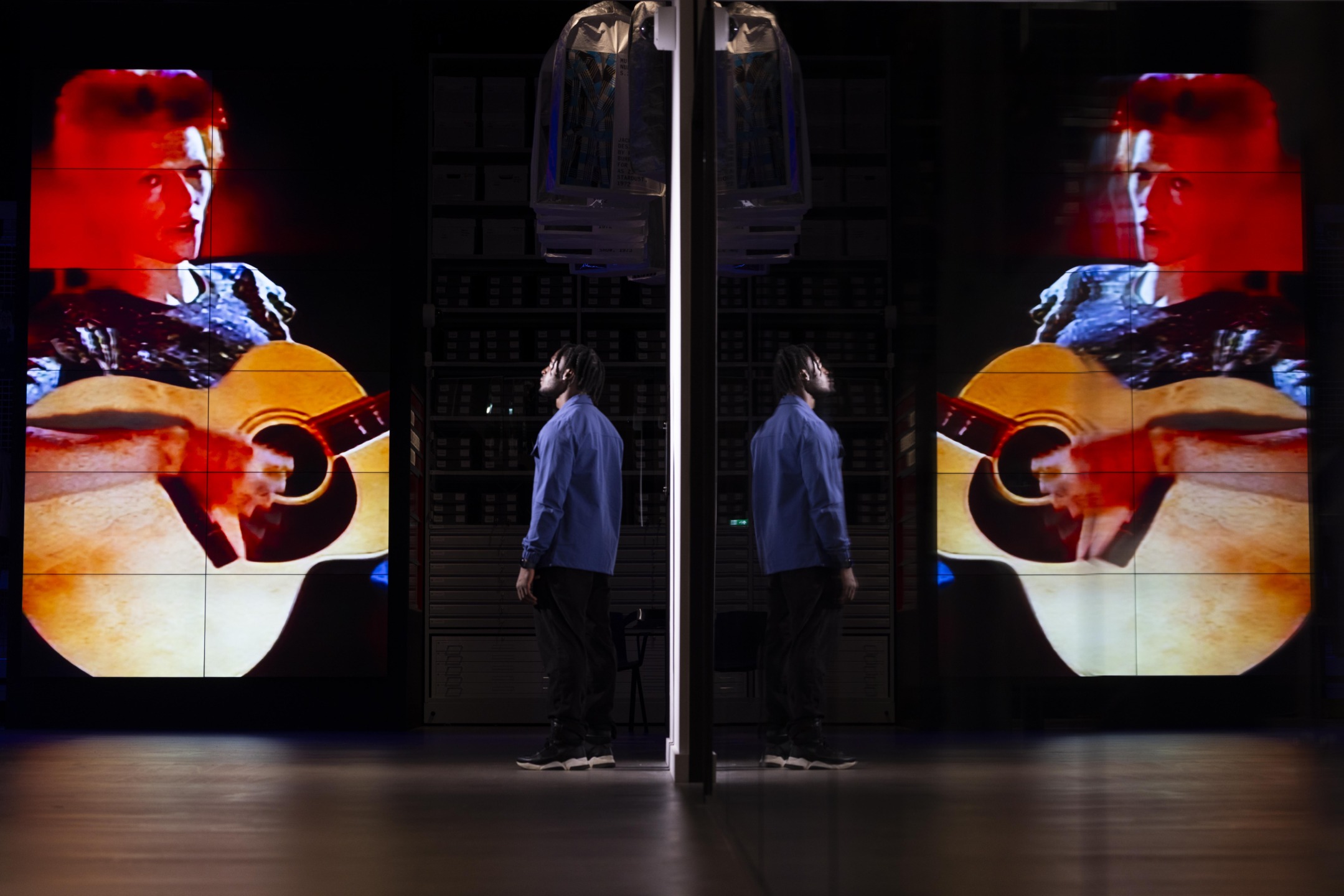 Person in blue looking at a large screen displaying a musician playing guitar, mirrored reflection on the surface.