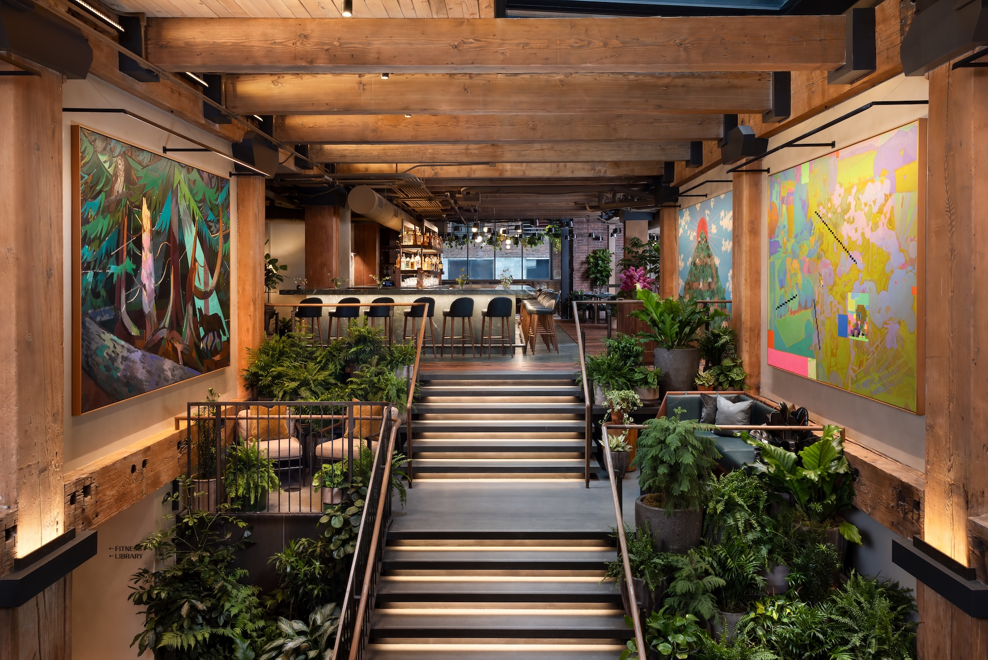 Modern interior with wooden beams, lush plants, and vibrant artwork, featuring a staircase leading to a bar area.