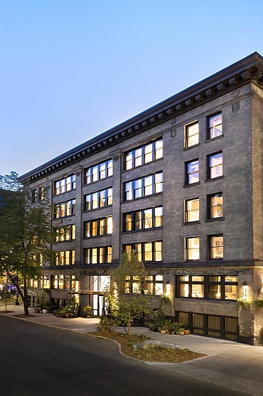 Five-story building with illuminated windows and a tree-lined street at dusk.