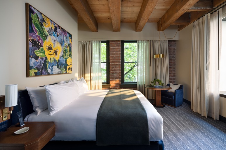 Cozy hotel room with a king-sized bed, colorful floral painting, wooden beams, and large windows letting in natural light.