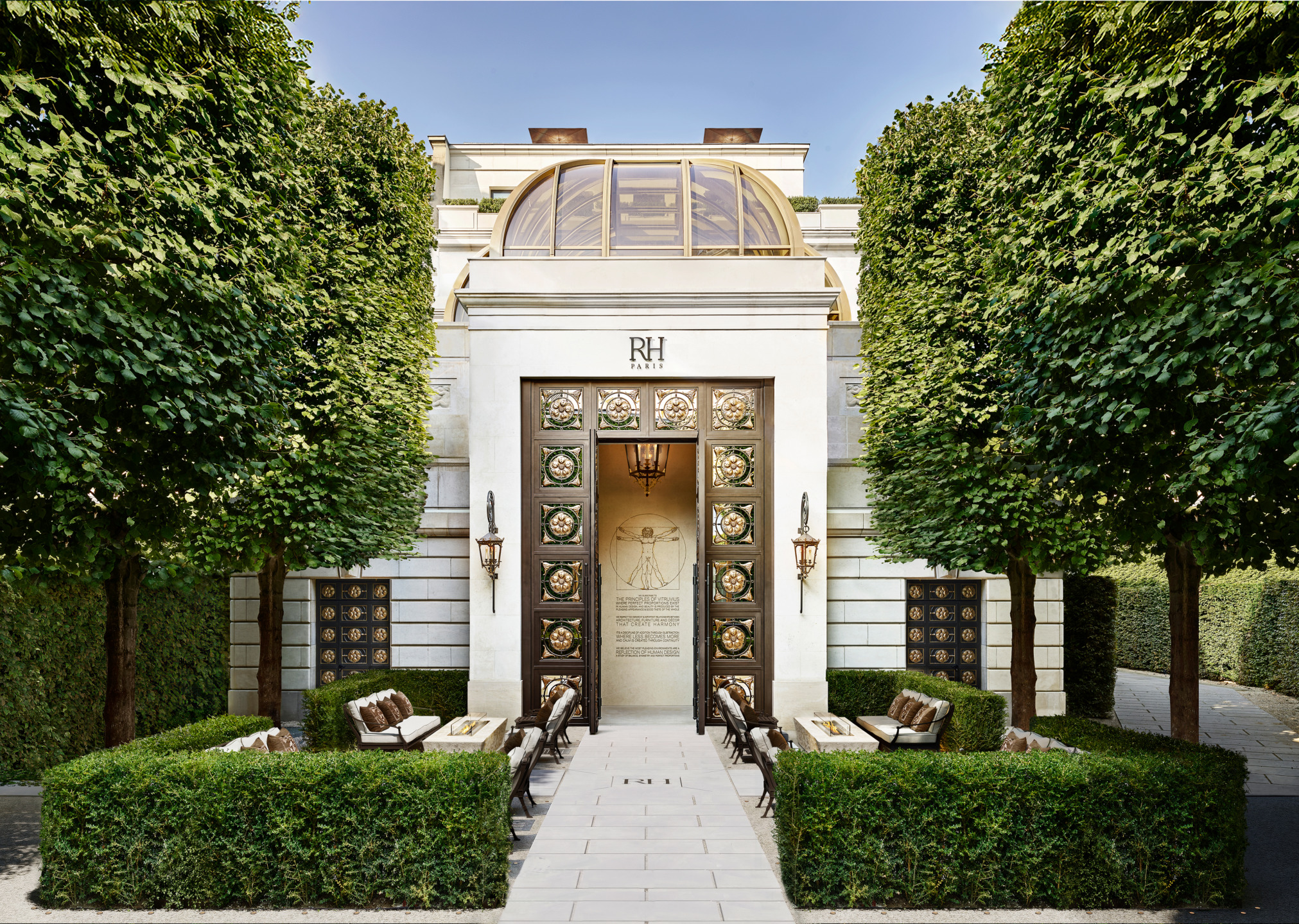 Elegant entrance to a luxury building with ornate doors, surrounded by manicured hedges and outdoor seating under tall trees.