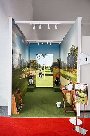 Indoor golf simulator setup with green grass, golf clubs, and a screen displaying a golf course scene.