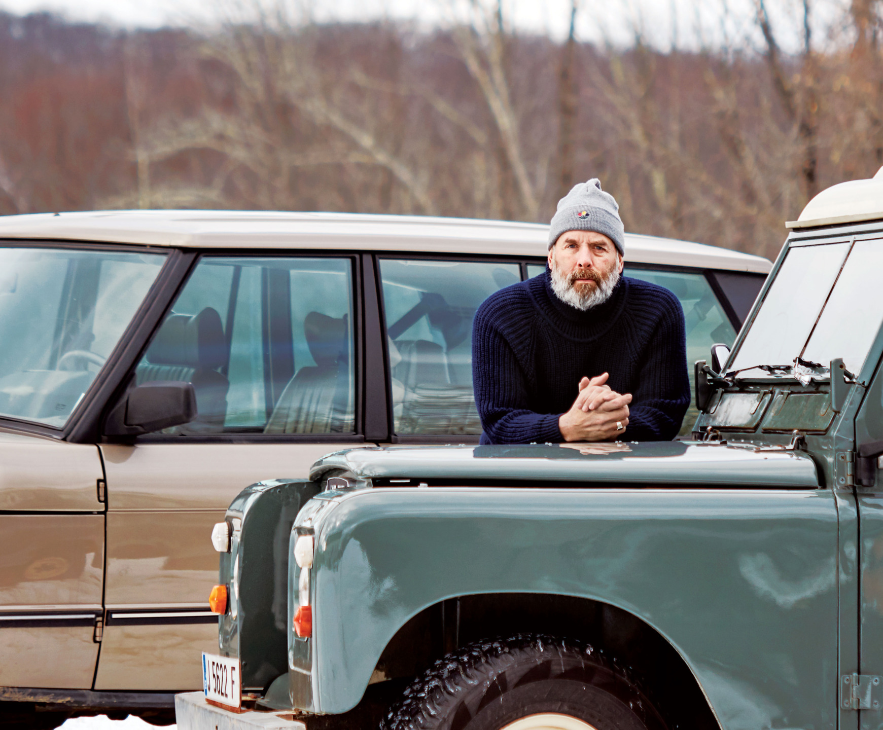 Man in a knit sweater and beanie leans on a vintage green vehicle parked next to a beige car in a rural setting.
