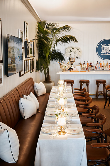Elegant restaurant interior with a long table set for dining, leather chairs, a bar, framed photos, and potted plants.