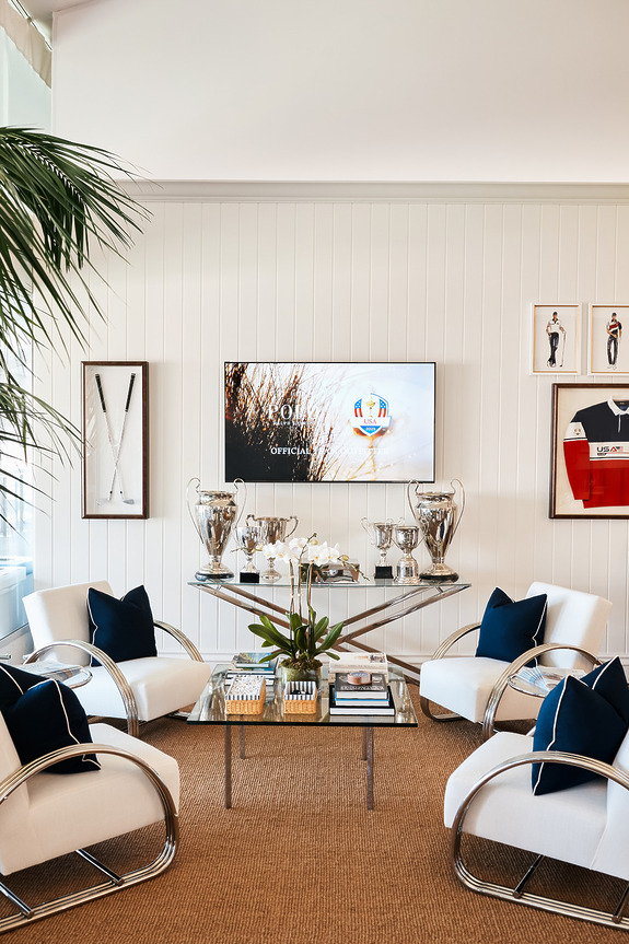 Luxurious living room with white armchairs around a glass table displaying elegant silver trophies and a wall-mounted TV.