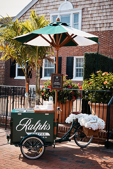 Green Ralph's Coffee bike cart with white flowers parked near a brick building, shaded by a large umbrella.