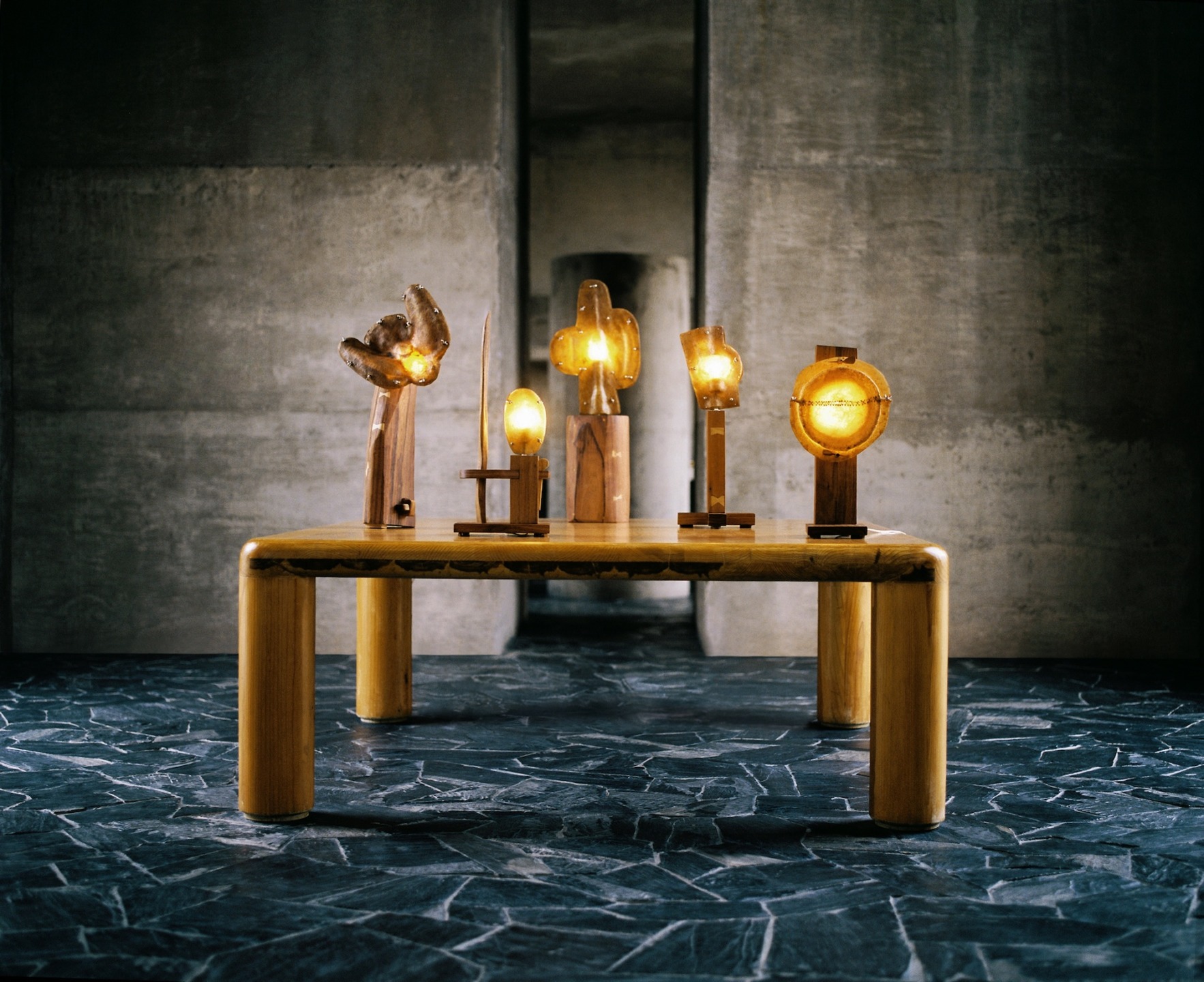 Wooden table with artistic wooden sculptures and glowing lamps on a dark textured floor in an industrial-style room.