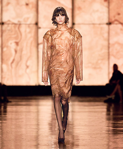 Model walking on a runway in a brown, translucent dress against a marble-patterned backdrop.