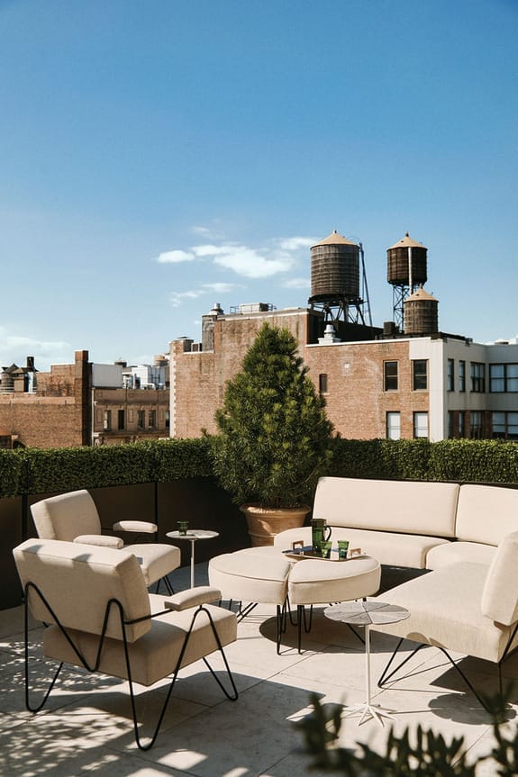 Outdoor rooftop patio with beige seating and tables, lush greenery, and a cityscape view featuring water towers against a clear sky