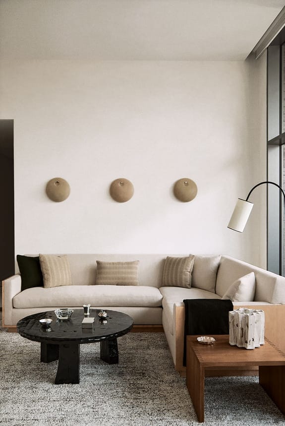 Modern living room with a beige sectional sofa, round black table, wooden side table, and wall-mounted decor pieces.