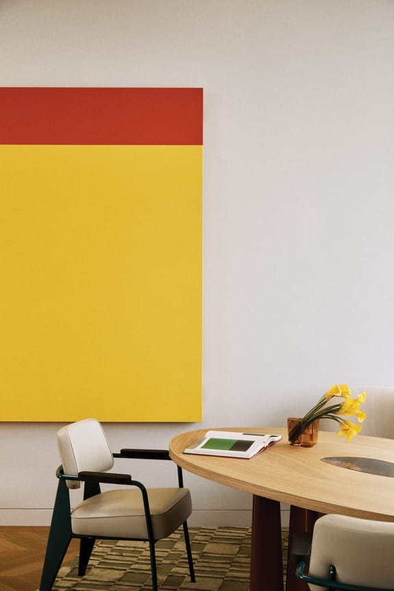Modern interior with a large yellow and red abstract painting, round wooden table, chairs, and flowers in a vase.