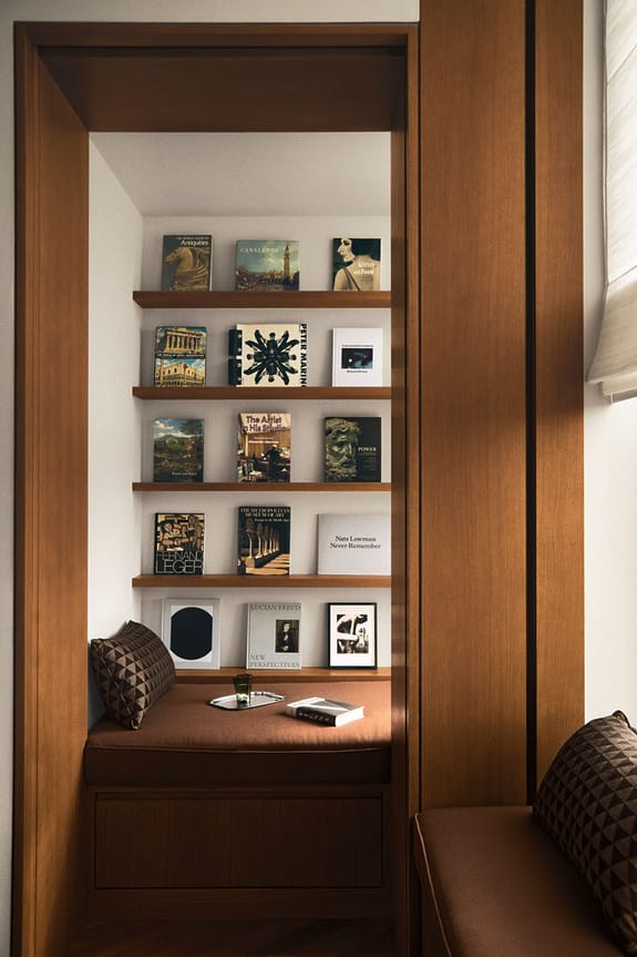 Cozy reading nook with wooden shelves displaying art books, a bench with cushions, and soft natural lighting.