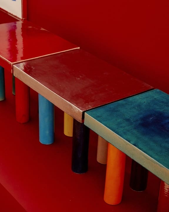 Colorful bench with shiny red, orange, and blue seats supported by cylindrical columns against a vibrant red wall.