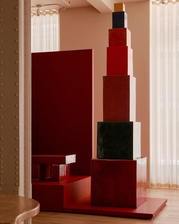 Stacked colorful blocks in a minimalist art installation with red and earthy tones in a contemporary room setting