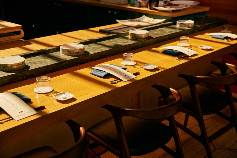 Elegant restaurant counter set for dining with plates, glasses, chopsticks, and napkins on a wooden surface in warm lighting.
