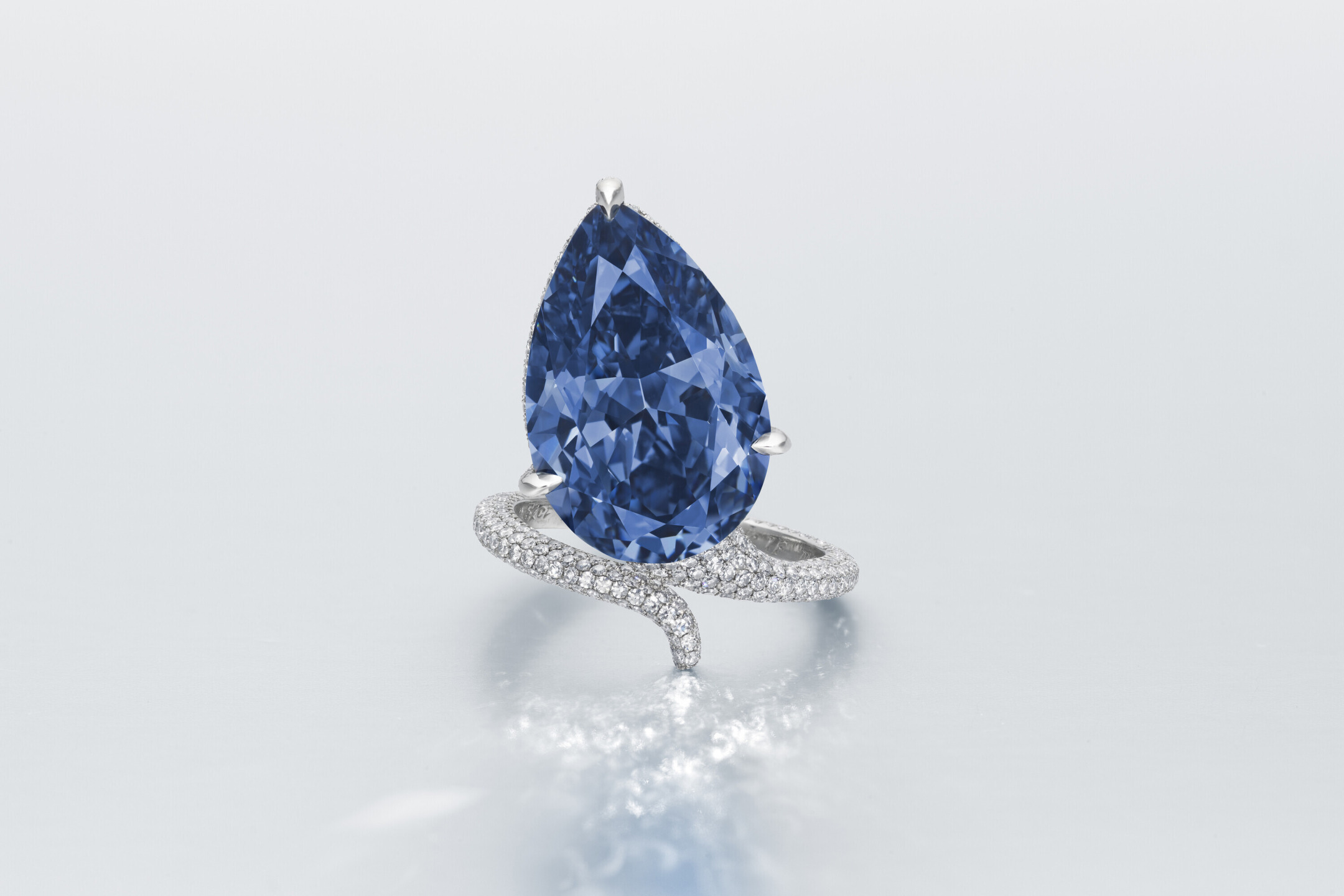 Pear-shaped blue diamond ring with a twisted band encrusted with small white diamonds on a reflective surface