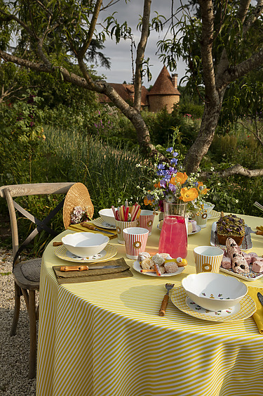Outdoor table setting with a yellow striped tablecloth, vibrant flowers, lemonade, and snacks under trees.