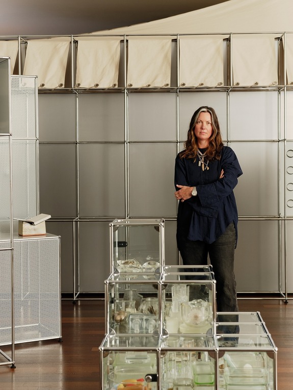 Person standing in a modern room with glass shelves and metal framing, looking at the camera.