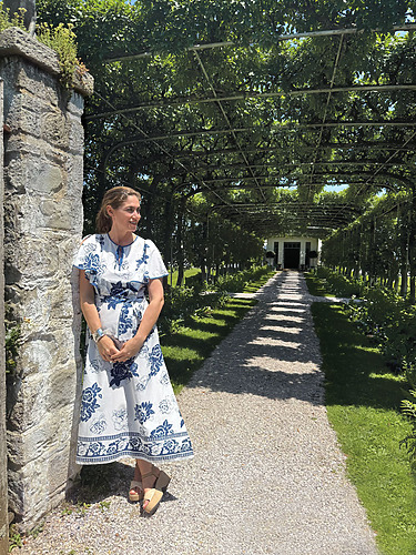 Woman in a white floral dress standing by a stone wall in a garden with a green archway path.