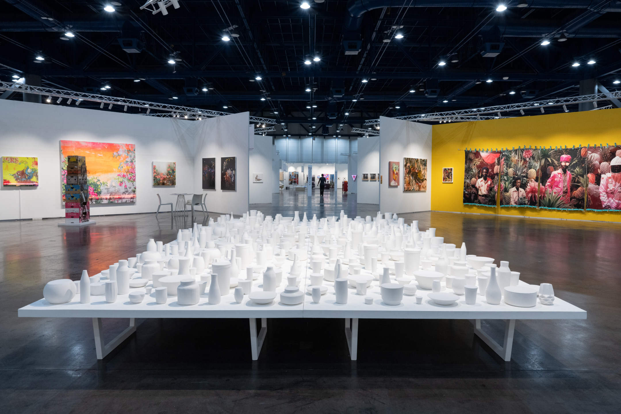 Art exhibition hall featuring a table with white sculptures, paintings on the walls, and spotlights illuminating the space.