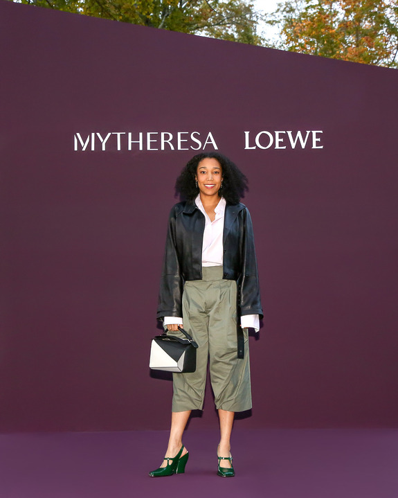 Person posing in front of a purple MyTheresa Loewe backdrop, wearing a stylish outfit with a handbag and green heels.