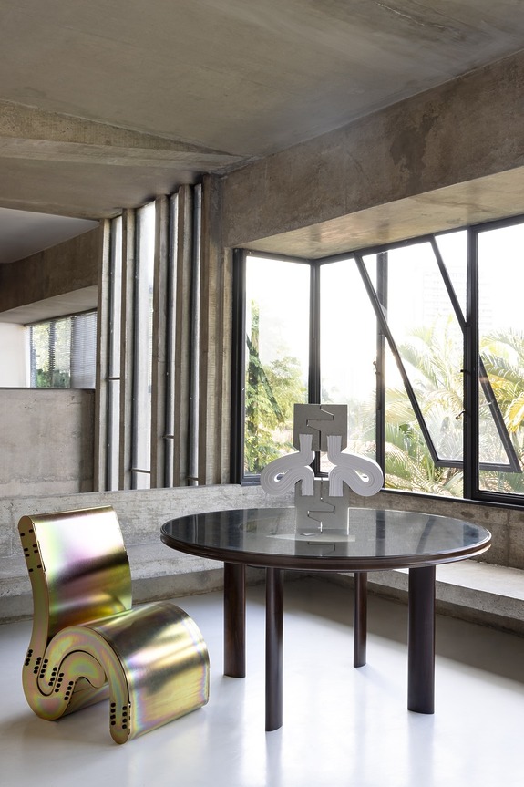 Modern interior with a glass table, abstract sculpture, and iridescent chair near large windows in a concrete room.