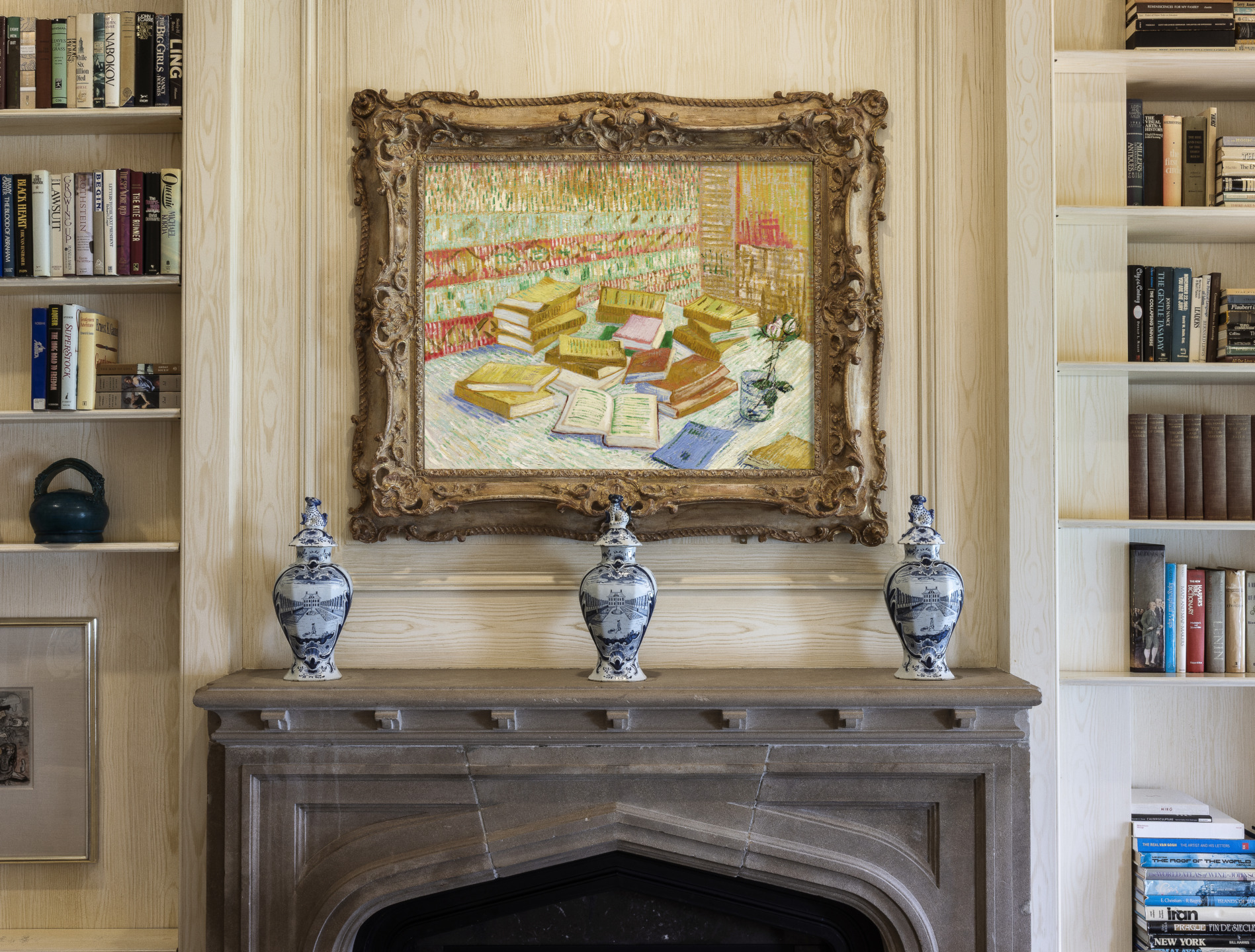 Framed painting of a library with bookshelves, hung above a mantel with three decorative vases, surrounded by bookshelves.