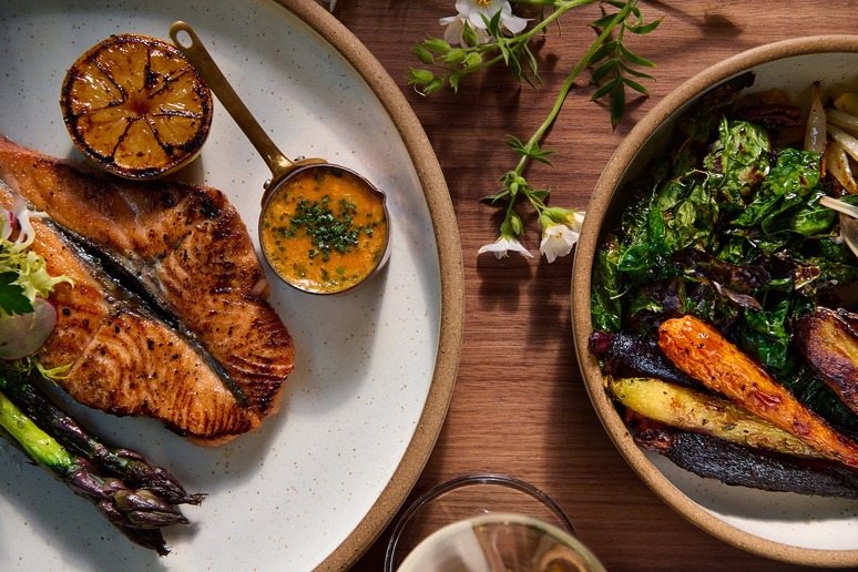 Grilled salmon with lemon, sauce, asparagus on one plate and roasted vegetables on another, garnished with flowers.
