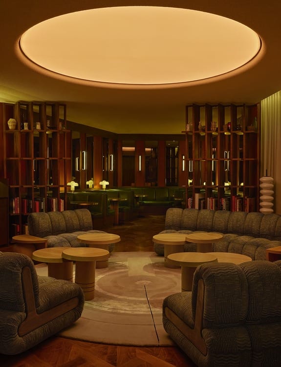 Cozy modern lounge with circular ceiling light, curved sofas, round tables, and warm ambient lighting.