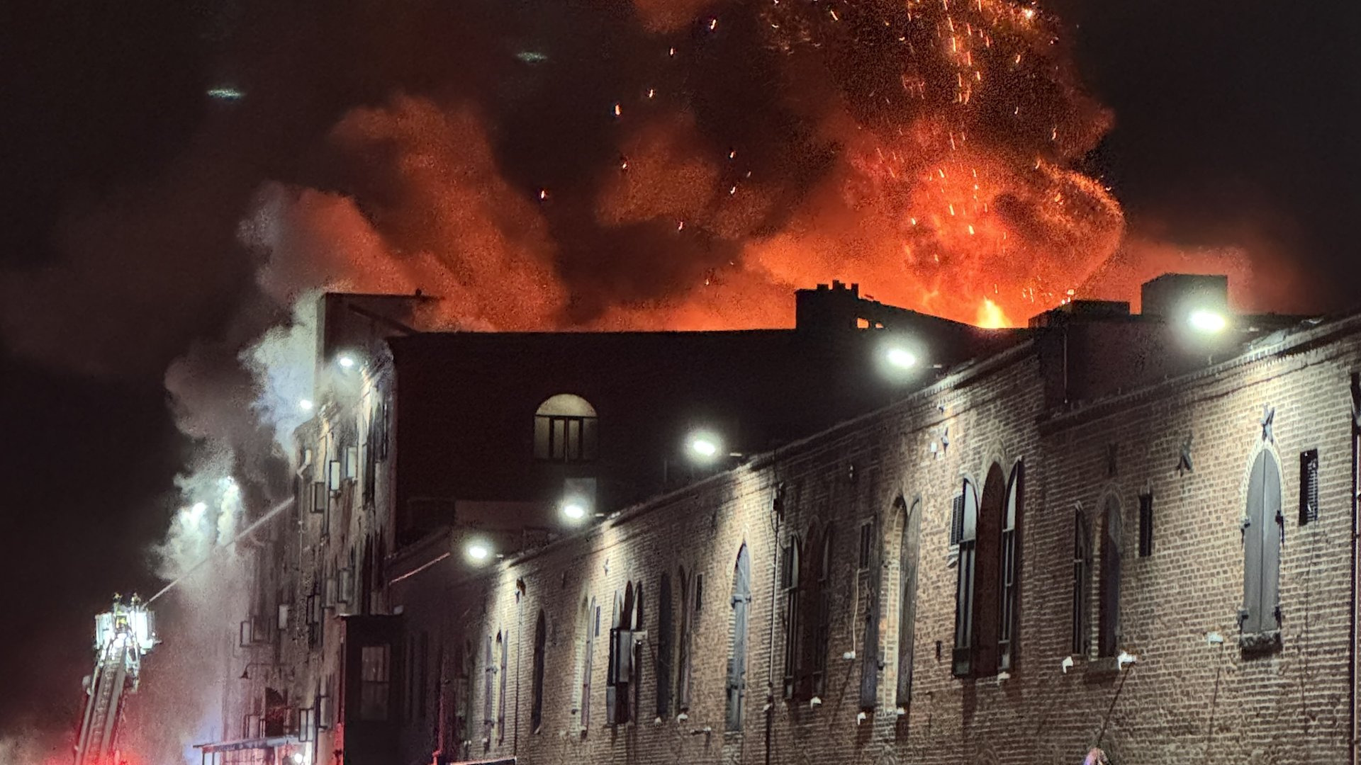Firefighters battle large blaze in a multistory building at night, with flames and heavy smoke rising from the roof.