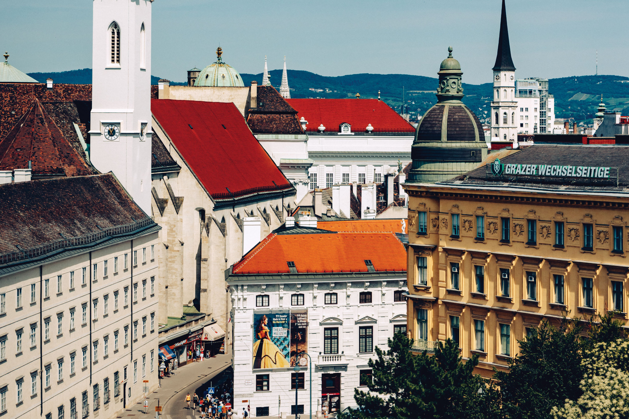 Scenic view of buildings with red roofs and a church tower in a European city against a backdrop of rolling hills.