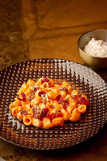 Pasta with creamy sauce and herbs on a textured plate, accompanied by a small bowl of white sauce on a wooden table.