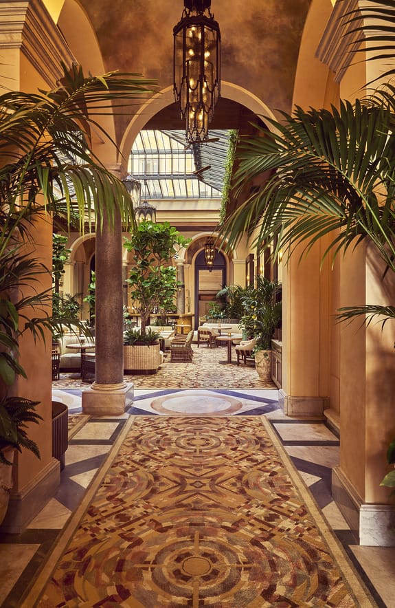 Elegant courtyard with ornate mosaic floor, tall columns, lush greenery, and a hanging chandelier creating a serene atmosphere.