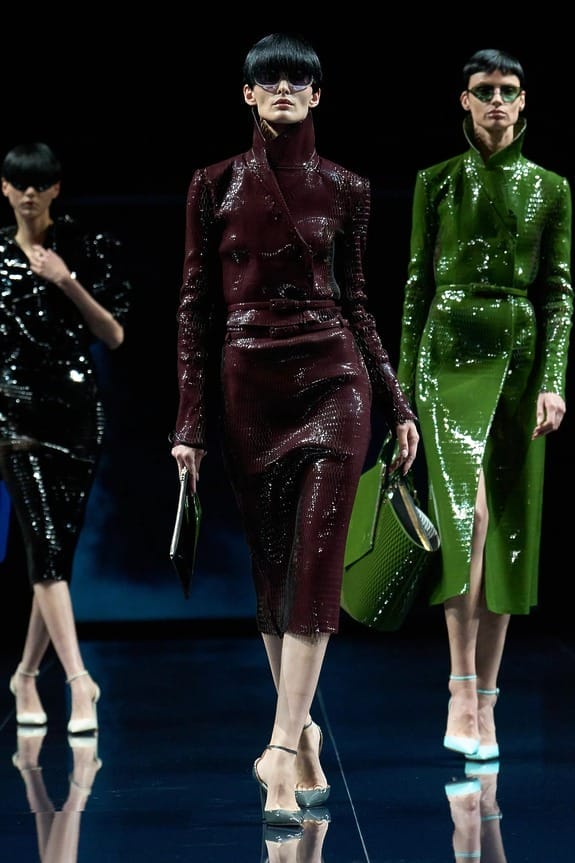 Models walking the runway in stylish, shiny outfits featuring dark red, black, and green designs with futuristic eyewear.